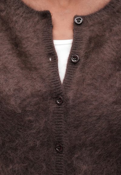 Cárdigan marrón esponjoso con cuello redondo y cinco botones oscuros, con una textura de punto y combinado con una camiseta blanca de debajo.