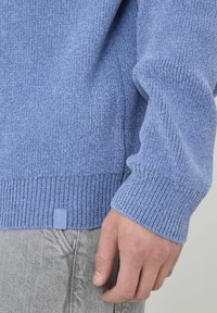 Sweater bleu clair avec un design en tricot texturé et des poignets côtelés. Petite étiquette logo visible à l'ourlet, associé à un pantalon gris.
