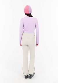 Svetlo fialový top s dlhými rukávmi v kombinácii s béžovými snehovými nohavicami, ktoré majú zipsy na členkoch a čierne lyžiarske topánky. Čiapka má ružové a fialové časti.