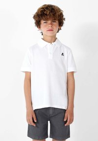 Camiseta polo blanca con un acabado texturizado y mangas cortas, que presenta un logo de calavera negra en el pecho, combinada con pantalones cortos gris oscuro.