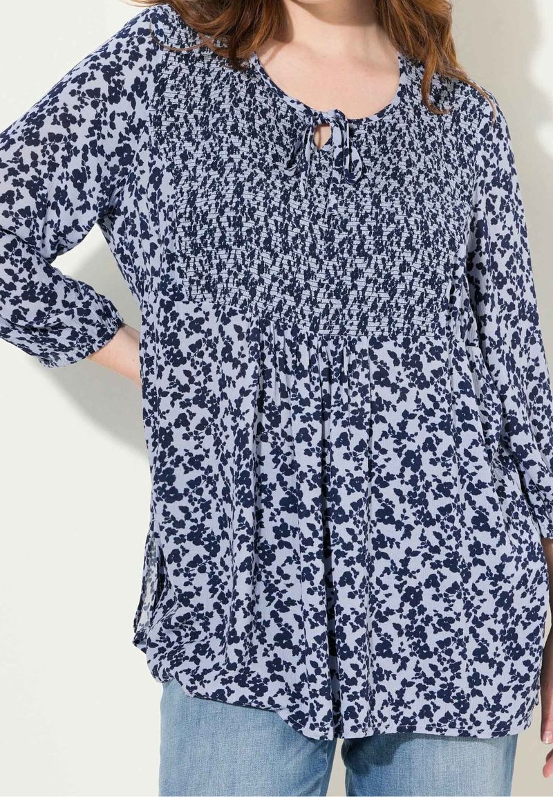Femme portant un chemisier ample à manches longues avec un motif floral bleu marine et un plissé au niveau de la poitrine, associé à un jean bleu clair.