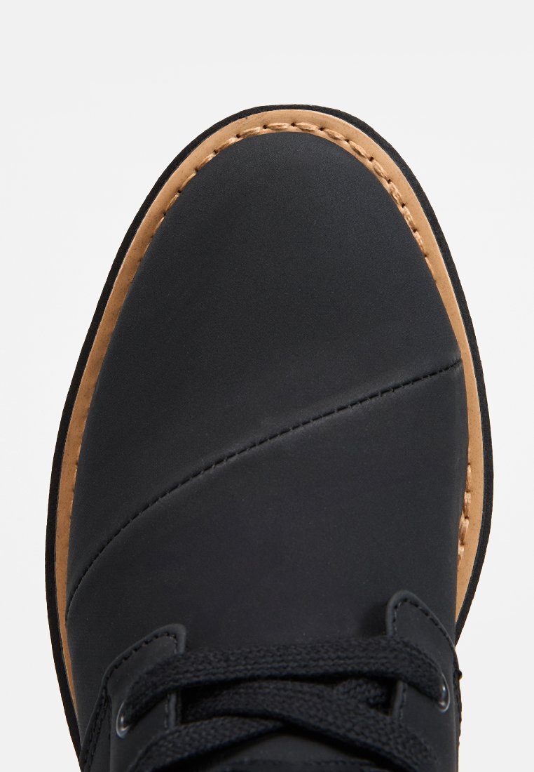 Czarny but z gładką skórzaną cholewką, brązowymi szwami, okrągłym noskiem i kontrastującą, beżową gumową podeszwą. Posiada czarne sznurowadła i oczka.