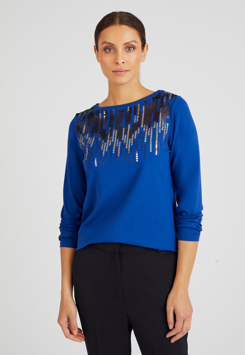 Top lungo a maniche blu con un motivo di paillettes nere lungo il collo, tessuto morbido e vestibilità rilassata. Abbinato a pantaloni scuri.