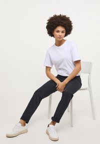 Fehér rövid ujjú póló, sötét farmernadrág és fehér sportcipő. A modell egy fehér széken ül, kezét a térdén nyugtatja, göndör haja van.