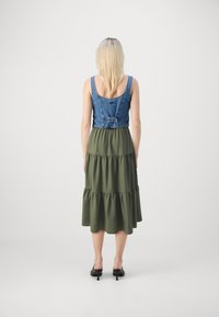 Cuerpo de denim con tirantes ajustables y falda verde de volantes. Textura suave, hasta la rodilla, combinada con mules negros de tacón.