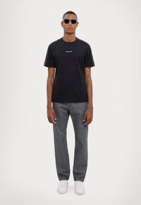 T-shirt en coton noir à manches courtes avec logo blanc. Associé à un jean en denim gris et des baskets blanches. Le modèle porte des lunettes de soleil noires.