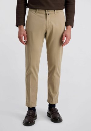 Beige Hosen mit geradem Schnitt, hergestellt aus leichtem Stoff. Mit einem Knopfverschluss und Taschen ausgestattet, kombiniert mit dunklen Schuhen und Socken.