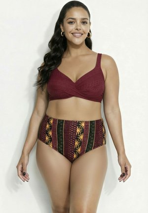 Kobieta z długimi ciemnymi włosami nosi bordowy, wiązany na zakładkę top od bikini oraz wysokie figi w wzory, uśmiechając się na jednolitym białym tle.
