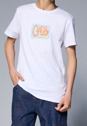 Person iført en hvid T-shirt med korte ærmer og et orange og blåt "GUESS Established in L.A." logo, kombineret med mørkeblå jeans.