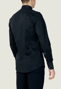 Maglietta nera a maniche lunghe con colletto alto, vestibilità su misura, texture liscia e polsini con bottoni. Orlo leggermente curvo e cuciture minime.