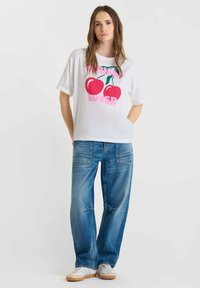 Femme debout, portant un t-shirt blanc avec des cerises rouges et un texte rose, associé à un jean bleu et des baskets blanches, sur fond uni.