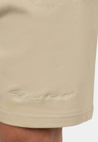 Pantaloni corti beige realizzati in tessuto morbido, con un logo ricamato in verde chiaro all'orlo, caratterizzati da una texture liscia e un design sartoriale.