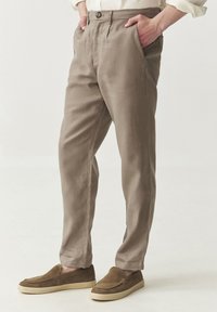 Beige Hosen mit gefaltetem front, Knopfverschluss und seitlichen Taschen, kombiniert mit braunen Mokassin-Schuhe aus strukturiertem Stoff.