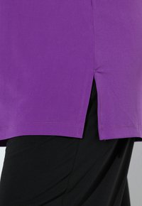 Fialová košile s krátkým rukávem, s rovým lemem a bočními rozparky, vyrobená z měkké tkaniny, vystavená vedle černých kalhot.