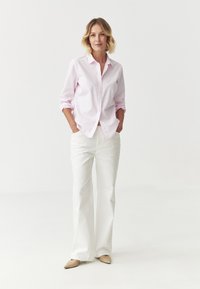 Chemise rose clair à boutons, manches retroussées, pantalon large blanc crémeux, et ballerines beiges à bout pointu. Design simple et épuré avec des couleurs neutres.