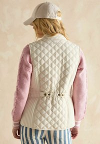 Gepolsterte cremefarbene Weste mit tailliertem Schnitt und Rautenmuster. Akzentuiert durch einen rosa gestrickten Ärmel und Knopfdetails in der Taille.