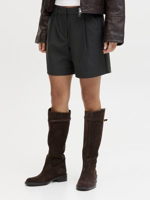 Person kledd i svarte plisserte shorts, mørkebrune knelangstøvler og en kort mørkebrunt skinnjakke på hvit bakgrunn.