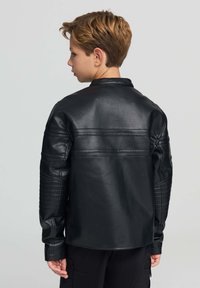 Ragazzo con capelli corti marroni che indossa una giacca di pelle nera e pantaloni neri, rivolto di lato su uno sfondo grigio uniforme.