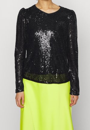 Personne portant un haut à manches longues noir à sequins assorti d'une jupe jaune fluo vif sur un fond clair uni.