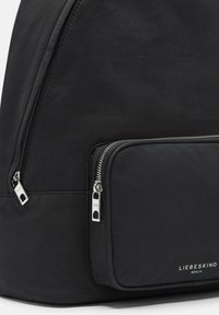 Zaino in tessuto nero con una grande tasca frontale con chiusura a zip e accessori in argento. Texture liscia con branding minimal.