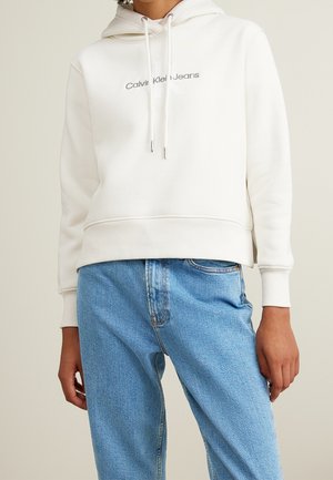 Personne portant un sweat à capuche blanc Calvin Klein Jeans avec cordons de serrage et un jean en denim bleu clair taille haute, debout devant un arrière-plan neutre.
