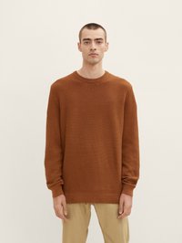Pulover texturat de culoare maro cu model canalat, decolteu rotund și mâneci lungi, asortat cu pantaloni bej deschis. Fundal simplu.