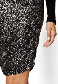 Falda negra con destellos plateados, que presenta un diseño fruncido y un corte a la altura de la rodilla. Textura suave con un brillo sutil. Mano visible en el encuadre.