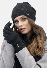Berretto in maglia nera con accenti di cristallo, guanti neri abbinati con polsini a coste e un cappotto grigio. Texture liscia con una vestibilità aderente.