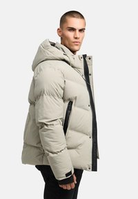 Fyllig jacka i ljusbeige, med huva, svarta dragkedjedetaljer och logotypaccent. Tillverkad med kviltad vaddering för isolering.
