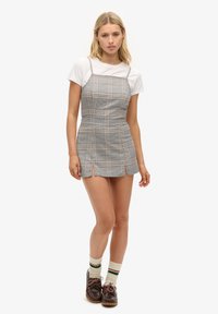 Junge Frau in einem weißen T-Shirt, das unter einem karierten Mini-Kleid mit Schlitz getragen wird, kombiniert mit gestreiften Socken und braunen Schnürschuhen, stehend vor weißem Hintergrund.