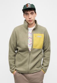 Νεαρός άνδρας που φοράει πράσινο καπέλο Salomon, ελιάς πράσινο φλις μπουφάν με κίτρινη τσέπη με φερμουάρ, πουκάμισο με σχέδια και μπεζ παντελόνι, με τα χέρια στις τσέπες.