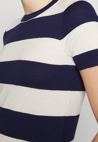 Pruhovaný pletený top s námořnickými modrými a krémovými horizontálními pruhy, krátkými rukávy a žebrovaným kulatým výstřihem s hladkou texturou.