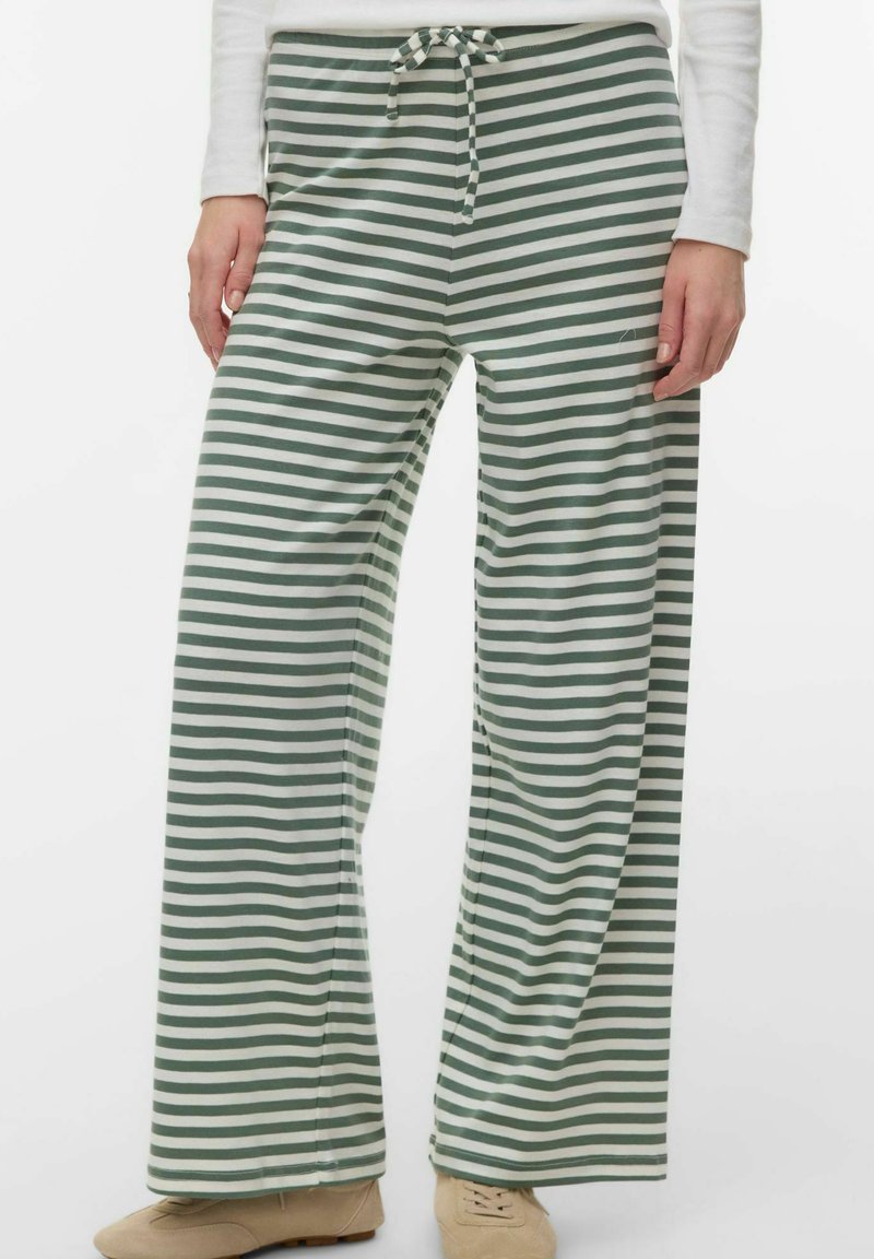 Partie inférieure du corps portant un pantalon large à rayures horizontales vertes et blanches avec une taille à cordon, et des chaussures beige à lacets.