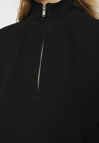 Černá mikina na zip s vysokým límcem, která má stříbrný kovový zip a hladkou, texturovanou látku.