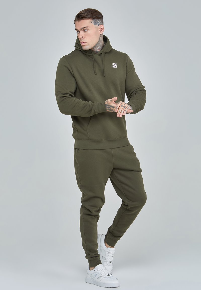 Conjunto de sudadera y pantalones jogger en color verde oliva, hecho de tela suave. La sudadera cuenta con un bolsillo frontal y un logo. Las zapatillas son blancas con costuras detalladas.