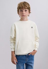Crème-kleurige sweatshirt met een ronde hals, een voorzak en een zwart schedel logo op de borst. G gedragen met blauwe spijkerbroek.