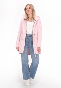 Világos rózsaszín esőkabát kapucnival, húzózsinóros díszítéssel, cipzáras záródással és oldalzsebekkel, kék, magas derekú farmert és fehér sportcipőt viselve.