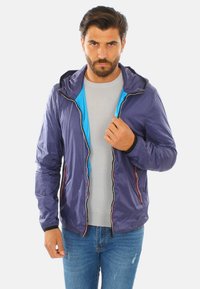 Giacca leggera viola con un interno blu a contrasto. Caratterizzata da un cappuccio, chiusura zip frontale e tasche con accenti rossi. Texture liscia.
