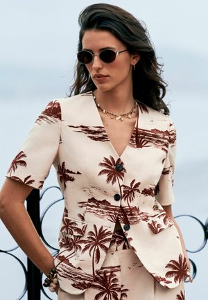 Kvinde iført beige kortærmet blazer og bukser med brunt palmetræsmønster, runde solbriller og lagdelte halskæder, udendørs ved gelænder.