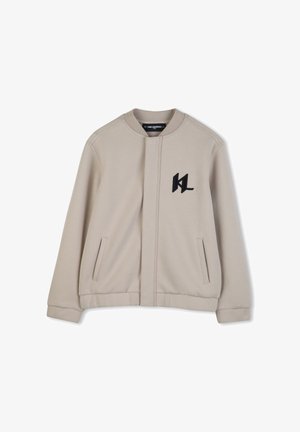 Giacca beige con zip in tessuto morbido, con scollo tondo, tasche laterali e logo nero ricamato sul petto a sinistra.