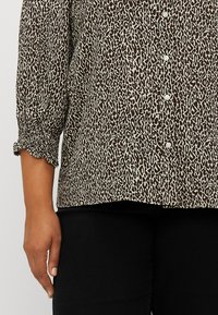 Blouse à imprimé léopard marron et crème avec des manches trois-quarts et des poignets froncés ; présente une rangée de boutons blancs sur le devant.