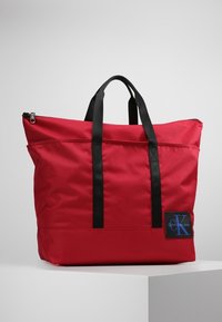 Borsa a tote rossa in tessuto con due manici neri, chiusura con zip e una toppa con logo blu. Texture liscia, forma rettangolare grande.