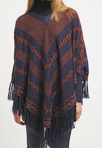 Marinblå och rostfärgad poncho med geometriskt mönster och fransade kanter, bärs över svart polotröja och byxor, täcker bålen och armarna.