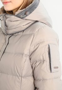 Mujer vestida con una chaqueta acolchada beige con capucha de piel sintética y bolsillo con cremallera en la parte superior de la manga, contra un fondo blanco.