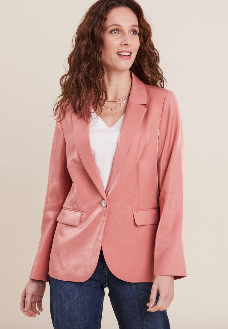 Blazer rosa in un tessuto liscio e lucido con chiusura a bottone singolo, due tasche frontali e un taglio sartoriale. Abbinato a un top bianco e jeans.