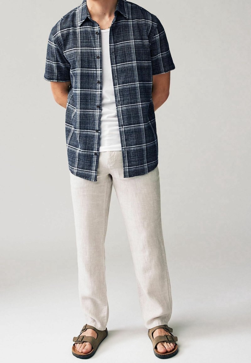 Camicia a maniche corte a quadretti blu navy con linee bianche, indossata sopra una canotta bianca. I pantaloni in lino beige e i sandali marroni completano l'outfit.