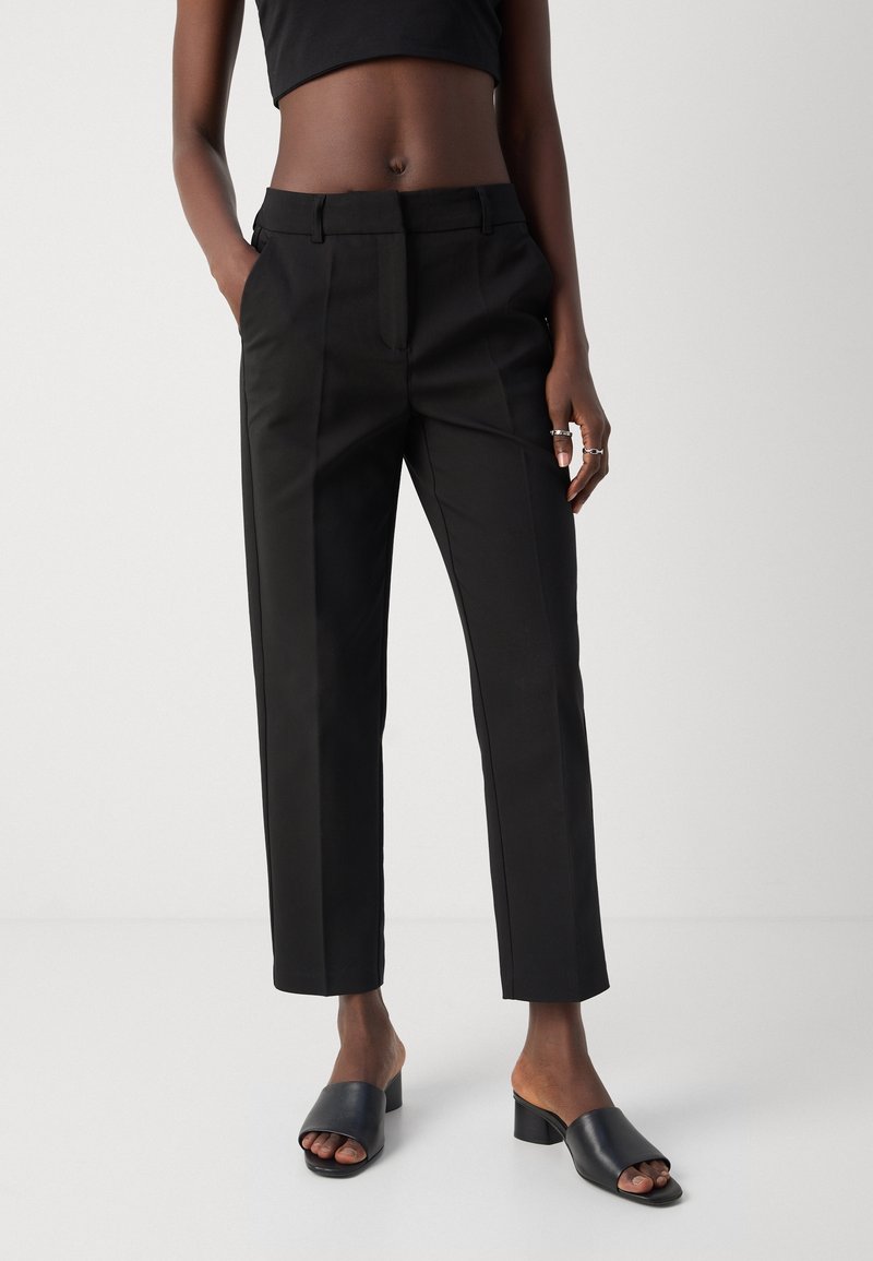 Pantalon noir slim sur mesure porté avec des mules noires à talons ouverts et un top court noir, mains dans les poches, sur fond uni.