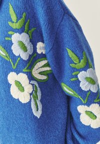 Pulover tricotat de culoare albastră, cu designuri florale brodate în alb și albastru deschis, cu frunzele și accente verzi, având o finisare texturată.