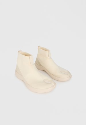 Paire de baskets beige montantes à la cheville avec orteil fendu, tissu élastique et semelles épaisses blanches sur fond clair.
