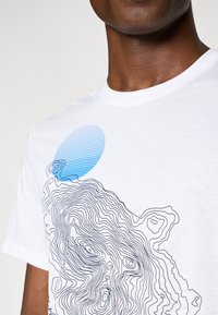 Hvid bomulds t-shirt med et sort konturdesign og en blå stribet cirkel, set fra halsområdet. Sømløs rund hals.